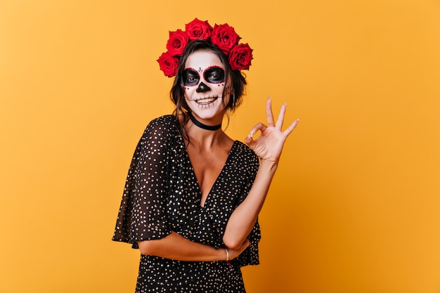 Gebräunte Frau mit schneeweißem Lächeln zeigt Zeichen ok. Foto der jungen Dame drinnen mit Halloween-Make-up auf orange Hintergrund.