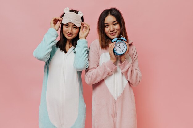 Gebräunte junge Frauen im Pyjama schauen nach vorne, lächeln und posieren auf rosa Wand