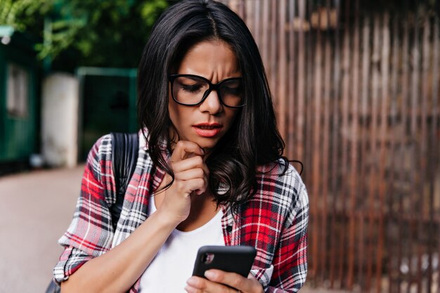 Gebräunte unglückliche Frau in den Gläsern, die auf der Straße mit Telefon stehen. Schönes ernstes Mädchen las Nachricht.