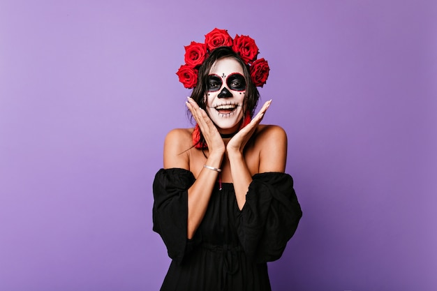 Gebräuntes Mädchen im schwarzen Kleid mit nackten Schultern in Überraschung. Innenporträt des jungen mexikanischen Modells mit Make-up für Halloween und Blumen in ihren Haaren