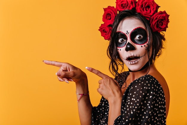 Gebräuntes weibliches Modell im Halloween-Outfit, das mit offenem Mund aufwirft. Wunderschönes Mädchen in traditioneller mexikanischer Kleidung, das den Tag der Toten feiert.