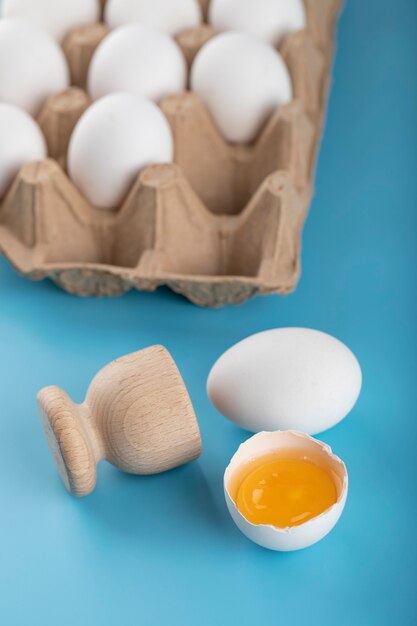 Gebrochenes rohes Ei und Behälter mit Eiern auf blauer Oberfläche.