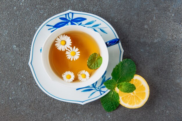 Gebrühter Kamillentee mit der Hälfte der Zitrone und der grünen Blätter in einer Tasse und Untertasse auf grauem Stuckhintergrund, Draufsicht.