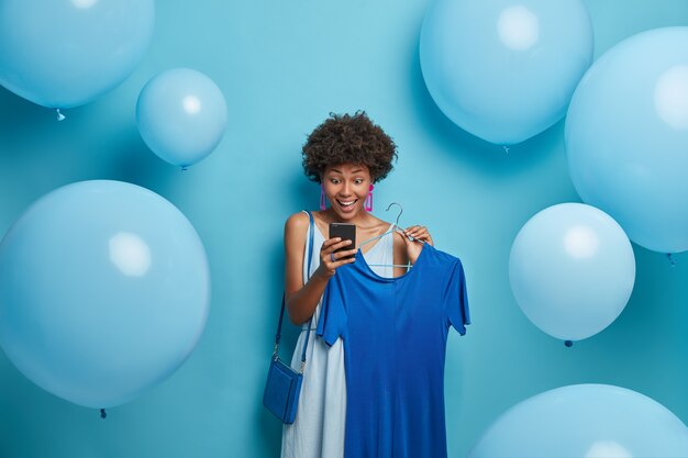 Geburtstag, Feiertage, Kleidungskonzept. Die freudig beeindruckte Frau starrt mit überraschtem, fröhlichem Blick auf das Smartphone-Display, erhält eine unerwartete Nachricht, nimmt ein Kleid auf Kleiderbügeln und kleidet sich in alles Blau