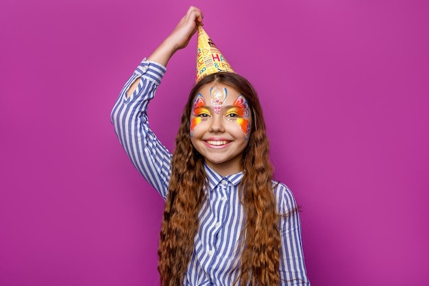 Geburtstagsmädchen mit bunten Faceart tragen Partymütze posiert isoliert auf violetter Wand.