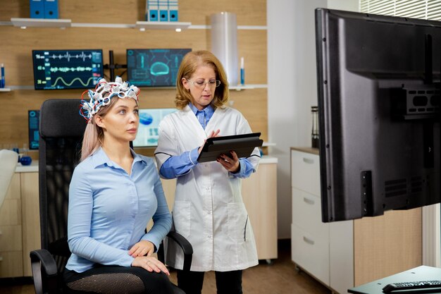 Geduldige Frau scannt ihr Gehirn und Arzt macht Notizen in der Tablette, die sie in der Hand hält. Gerät zum Scannen von Gehirnwellen