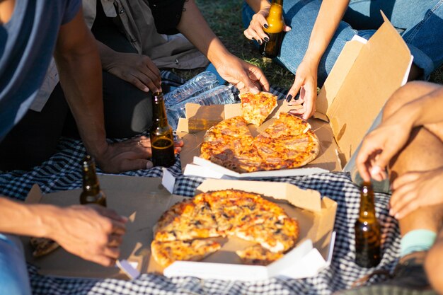 Geernteter Schuss von den Freunden, die Picknick im Sommerpark haben. Junge Leute sitzen auf der Wiese mit Pizza und Bier. Konzept des Picknicks