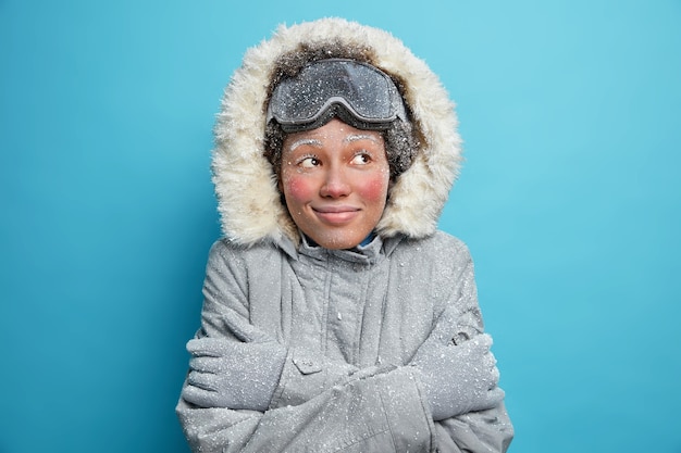 Gefrorene Frau umarmt sich zitternd vor Kälte bedeckt mit Raureifblicken trägt gerne Snowboardbrille Winterjacke und Handschuhe lächelt angenehm.