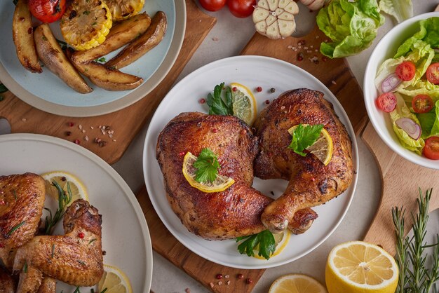 Gegrillte Hähnchenschenkel in Barbecue-Sauce und geröstetem Gemüse und gemischtem Gemüsesalat mit Tomaten, Zitrone in weißer Platte auf hellem Steintisch.