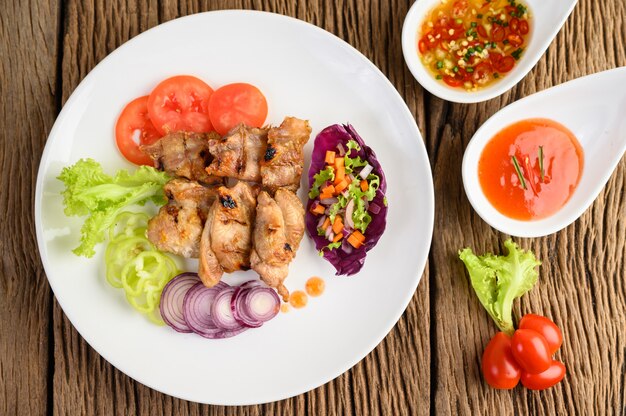 Gegrilltes Hähnchen auf einem weißen Teller mit Salat, Tomaten, in Stücke geschnittenen Chilis und Sauce auf Holztisch.