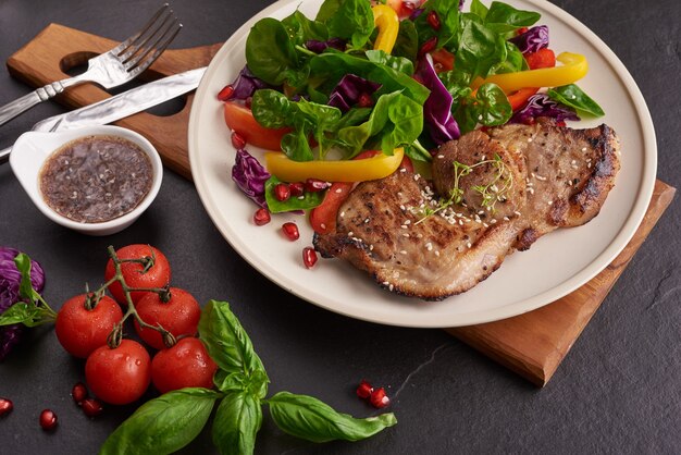 Gegrilltes Steak mit frischem Gemüse, Paprika, Tomaten, roten Zwiebeln, rosa Pfeffer und Gewürzen. Hausgemachtes leckeres Essen. Konzept für leckeres und gesundes Essen. Schwarze Steinoberfläche. Schweinesteak mit Salat