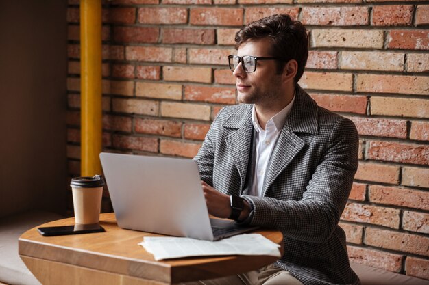 Geheimnisgeschäftsmann in den Brillen, die durch die Tabelle im Café mit Laptop-Computer sitzen und weg schauen