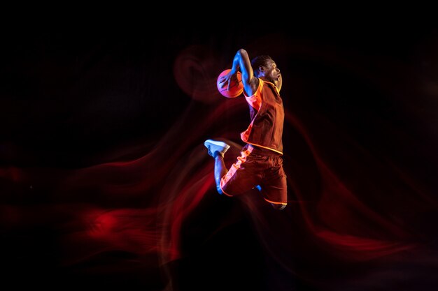 Geheimnisvolle Natur. Afroamerikanischer junger Basketballspieler des roten Teams in Aktion und Neonlichtern über dunklem Studiohintergrund. Konzept von Sport, Bewegung, Energie und dynamischem, gesundem Lebensstil.