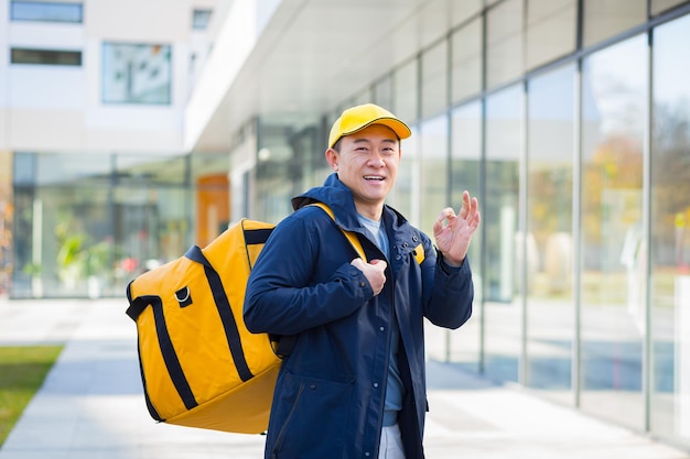 Gehender kurierlieferant, der asiatisch lächelt und in die kamera schaut, hat einen großen gelben rucksack für die lebensmittellieferung
