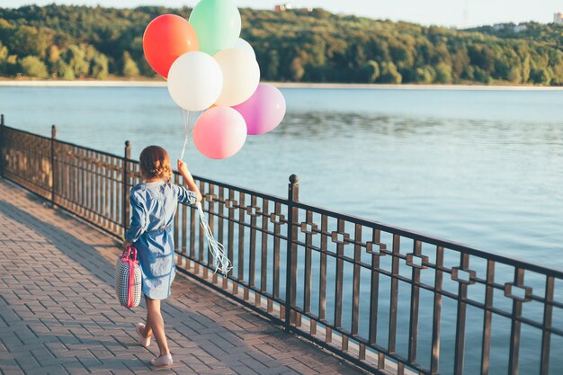 Gehendes Mädchen, das bunte Luftballons und kindlichen Koffer hält