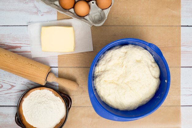 Gekneteter Teig in blauer Schale; Eier; Butter; Mehl; und Nudelholz auf Schreibtisch aus Holz