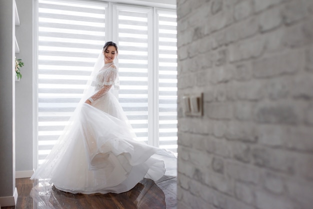 Gelächelte hübsche Braut dreht sich im Zimmer nahe weißer Backsteinmauer um, gekleidet in modisches Kleid, Hochzeitsmode