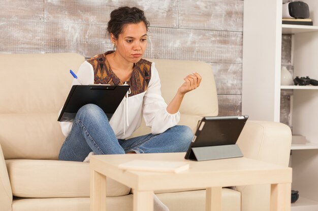 Gelangweilte Freiberuflerin bei der Arbeit von zu Hause aus und schaut auf den Tablet-Computer, während sie Notizen macht.