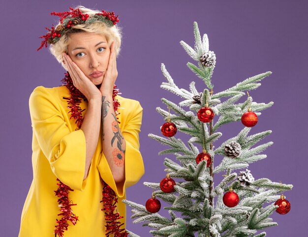 Gelangweilte junge blonde Frau, die den Weihnachtskopfkranz und die Lametta-Girlande um den Hals trägt, die in der Nähe des verzierten Weihnachtsbaums stehen und die Kamera betrachten, die Hände auf Gesicht lokalisiert auf lila Hintergrund hält