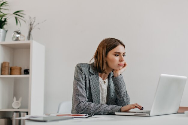 Gelangweilte junge Geschäftsdame im grauen Outfit schaut auf Laptop-Bildschirm an ihrem Arbeitsplatz.