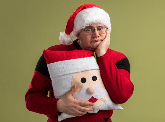 Gelangweilter erwachsener Mann mit Brille und Weihnachtsmütze, der das Weihnachtsmann-Kissen hält und die Hand auf dem Gesicht hält und die Seite isoliert auf olivgrüner Wand betrachtet