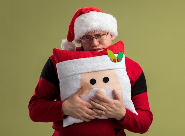 Kostenloses Foto gelangweilter erwachsener mann mit brille und weihnachtsmütze, der ein weihnachtsmann-kissen von hinten hält, isoliert auf olivgrüner wand?