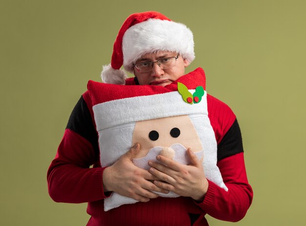 Gelangweilter erwachsener Mann mit Brille und Weihnachtsmütze, der ein Weihnachtsmann-Kissen von hinten hält, isoliert auf olivgrüner Wand?