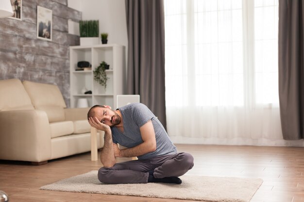 Gelangweilter Mann, der während der Selbstisolation auf Teppich im Wohnzimmer sitzt.
