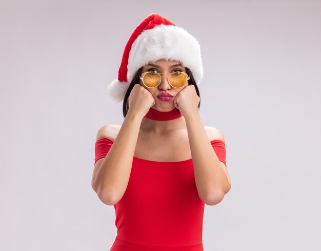 Gelangweiltes junges Mädchen mit Weihnachtsmütze und Brille, das in die Kamera schaut und die Hände auf dem Gesicht hält, mit geschürzten Lippen isoliert auf weißem Hintergrund mit Kopierraum