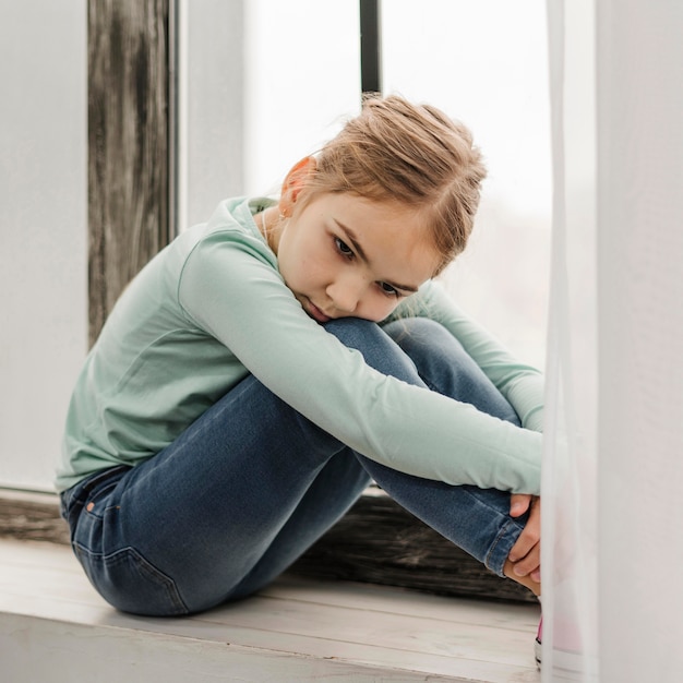 Kostenloses Foto gelangweiltes kleines mädchen, das auf einem fensterbrett sitzt