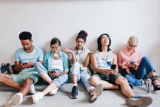 Gelangweiltes Mädchen mit lockigen schwarzen Haaren wütend, weil ihre College-Freunde ihr nicht zuhören. Müde internationale Studenten sitzen auf dem Boden und benutzen diese Telefone nach der Vorlesung.