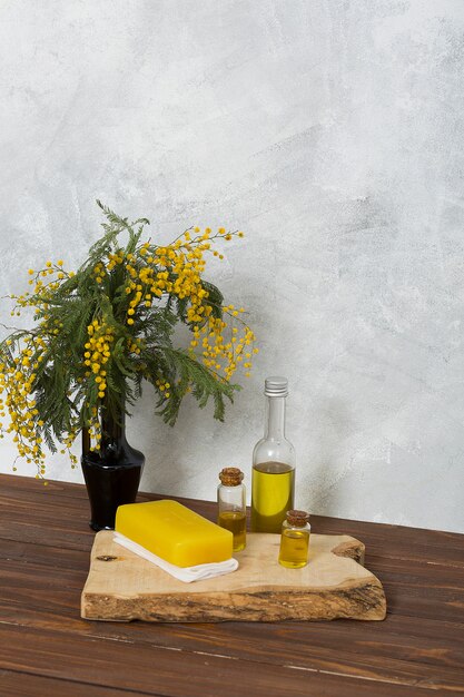 Gelber Mimosenblumenvase mit Kräuterseife und Flasche des ätherischen Öls auf hölzernem Brett über Tabelle gegen graue Wand