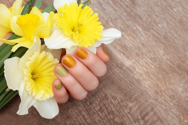 Gelbes Nageldesign. Weibliche Hand mit Glitzer-Maniküre, die Narzissenblüten hält.