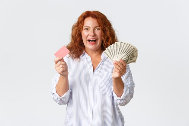 Geld-, Finanz- und Personenkonzept. Fröhliche und aufgeregte rothaarige Frau mittleren Alters in lässiger Bluse, die Geld und Kreditkarte mit optimistischem Lächeln hält, weißen Hintergrund stehend.