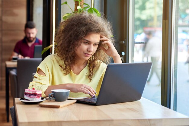 Gelockter Student, der mit Laptop arbeitet, sitzt im Café