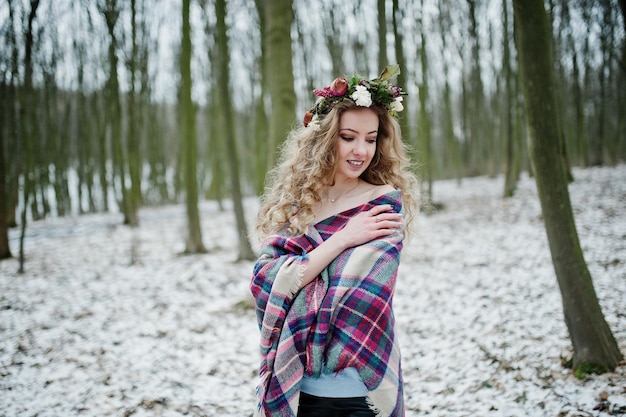 Gelocktes süßes blondes Mädchen mit Kranz im karierten Plaid am verschneiten Wald am Wintertag