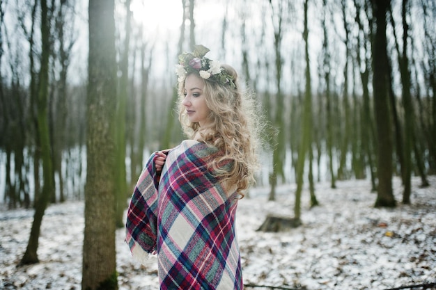 Gelocktes süßes blondes Mädchen mit Kranz im karierten Plaid am verschneiten Wald am Wintertag