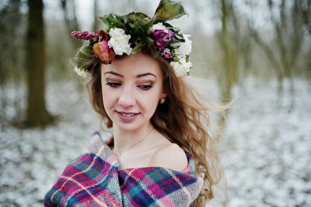 Gelocktes süßes blondes Mädchen mit Kranz im karierten Plaid am verschneiten Wald am Wintertag