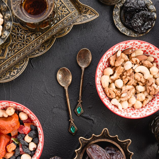 Gemischte Nüsse; Tee; Trockenfrüchte und metallische Löffel auf schwarzem konkretem Hintergrund