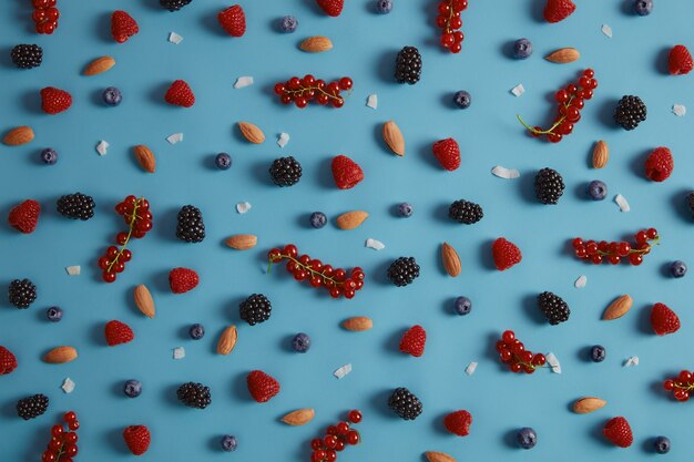Gemischte sortierte frische Beeren einschließlich Himbeere, Blaubeere, Brombeere, rote Johannisbeere auf blauem Hintergrund. Mandelnüsse und Kokosflocken. Gesunde Zutaten für ein leckeres Dessert. Große Vielfalt