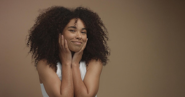 Gemischtes Rennen schwarzes Frauenporträt mit großem Afro-Haar, lockiges Haar auf beigem Hintergrund, das ihren Skating glücklich berührt