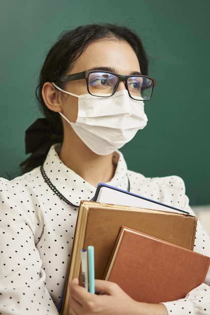 Gemischtrassige lehrerin, die eine gesichtsmaske trägt und ihre lehrbücher in einem klassenzimmer hält covid19