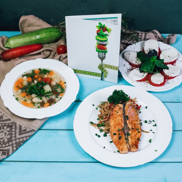 Gemüsesuppe, gegrilltes Fischfilet und Mozarellasalat auf dem blauen Holztisch.