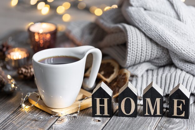 Gemütliche Winterkomposition mit einer Tasse Tee, dem dekorativen Wort Zuhause, Strickelementen und Bokeh-Lichtern.