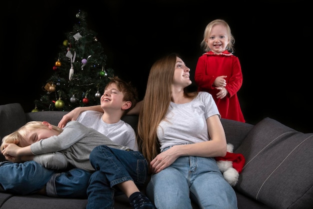 Gemütlicher familienabend - mama und drei glücklich lächelnde kinder vor dem hintergrund des weihnachtsbaums.