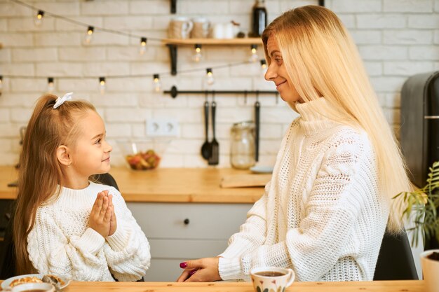 Gemütliches Bild des verblüfften entzückenden kleinen Mädchens mit aufgeregtem Blick, der am Küchentisch mit ihrer Mutter kocht oder frühstückt, Tee trinkt, warme Pullover trägt. Gemütliche festliche Atmosphäre