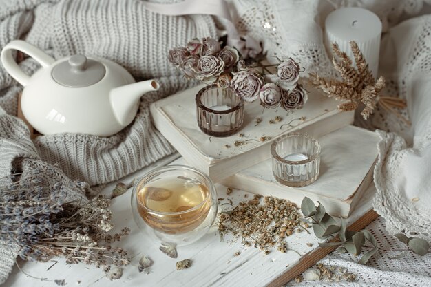 Gemütliches Lichtstillleben mit Kerzen, einer Tasse Tee, einer Teekanne und trockenen Kräutern.
