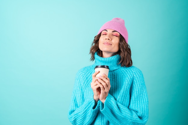 Gemütliches Porträt einer jungen Frau in einem gestrickten blauen Pullover und einer rosa Mütze mit hellem Make-up, die eine Tasse heißen Kaffee hält