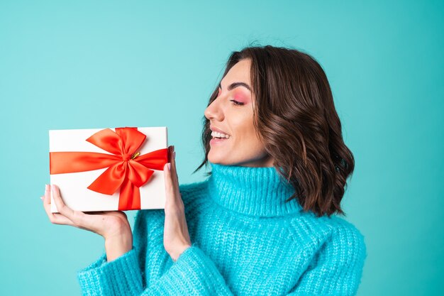 Gemütliches Porträt einer jungen Frau in einem gestrickten blauen Pullover und leuchtend rosa Make-up mit einer Geschenkbox