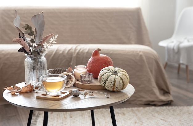 Gemütliches Wohnstilleben mit einer Tasse Tee, Kürbissen, Kerzen und Herbstdekordetails auf einem Tisch auf einem unscharfen Hintergrund des Raumes.